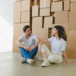 A person sitting on the sidewalk outside an apartment building surrounded by moving boxes, representing the millions of working Americans losing their homes to rent increases and evictions in America's 2026 housing crisis