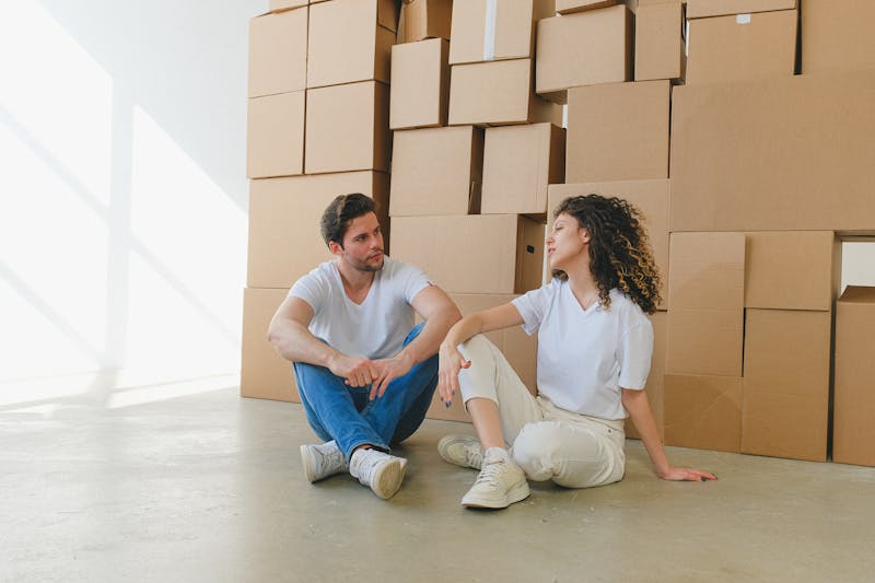 A person sitting on the sidewalk outside an apartment building surrounded by moving boxes, representing the millions of working Americans losing their homes to rent increases and evictions in America's 2026 housing crisis